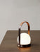A close-up of the Carrie Table Lamp in bronzed brass, its leather-wrapped handle and lit opal glass shade on a wooden table.