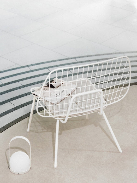 A white Carrie Table Lamp sits on a light grey floor beside a white wireframe String Chair with a magazine on its seat.