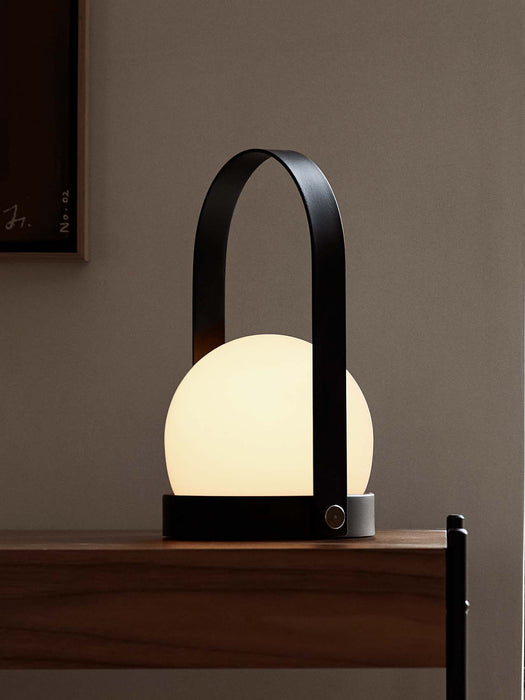 A close-up of the black portable Carrie Table Lamp, its white orb glowing warmly on a dark wood shelf against a taupe wall.