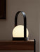 A close-up of the black portable Carrie Table Lamp, its white orb glowing warmly on a dark wood shelf against a taupe wall.