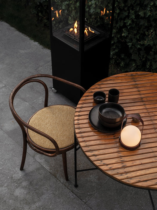 A lit Carrie Table Lamp on a wooden patio table with black dinnerware, next to a bentwood chair and an outdoor fireplace.