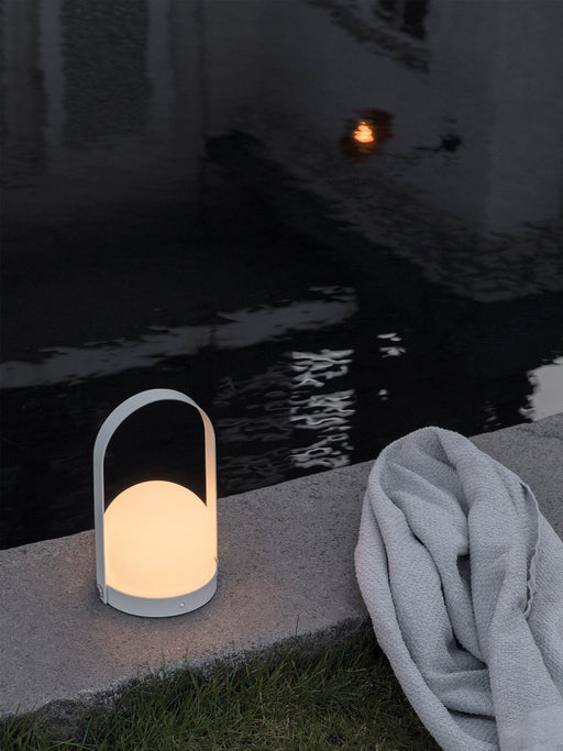 A white, illuminated Carrie Table Lamp rests on the stone edge of a swimming pool at night, a light grey towel next to it.