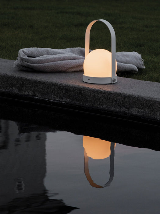 A glowing white Carrie Table Lamp sits on a stone ledge by the water at dusk, its reflection visible next to a grey towel.