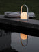 A glowing white Carrie Table Lamp sits on a stone ledge by the water at dusk, its reflection visible next to a grey towel.