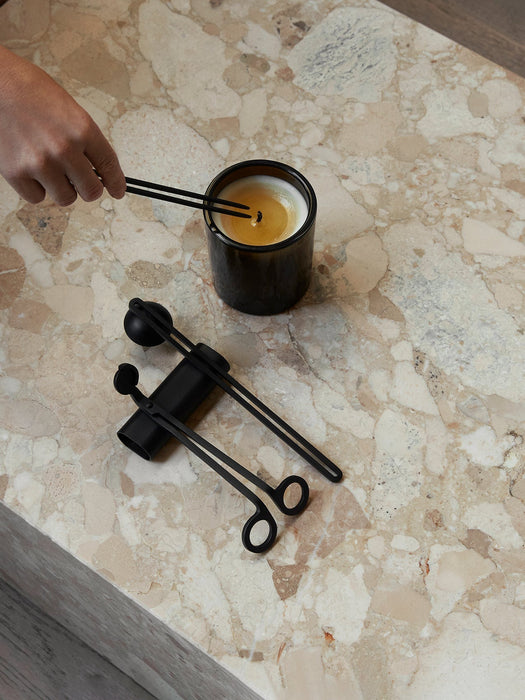 A hand tends a candle with the Audo Candle Care Kit wick dipper. The trimmer and snuffer rest nearby on a marble surface.