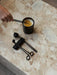 A hand tends a candle with the Audo Candle Care Kit wick dipper. The trimmer and snuffer rest nearby on a marble surface.