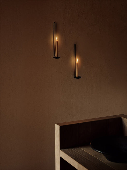 Two minimalist black Clip Candle Holder Wall sconces from Audo CPH, their lit candles casting a warm glow on a brown wall.