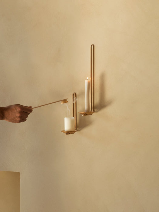 Two brass Clip Candle Holder Wall on a beige wall. A hand uses a snuffer to extinguish one candle, while the other is lit.