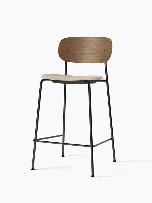 An angled view of the Co Bar Chair, featuring a dark stained oak backrest, a light bouclé seat, and a black steel frame.