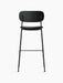 A tall, black Co Bar Chair from behind, with its black oak backrest, upholstered seat, and minimalist steel frame.