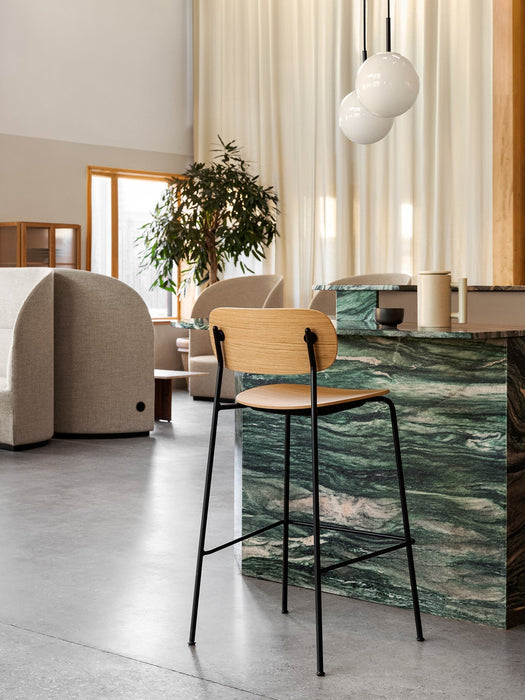 A Co Bar Chair in oak at a green marble bar. In the background are a Tearoom Sofa, Lounge Chair, and a potted plant.