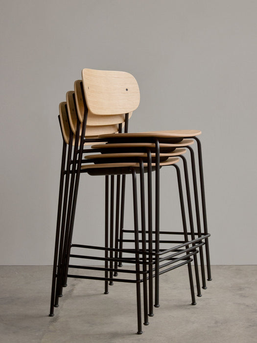 A stack of four Co Bar Chairs in natural oak veneer with black steel frames, shown against a plain grey wall.