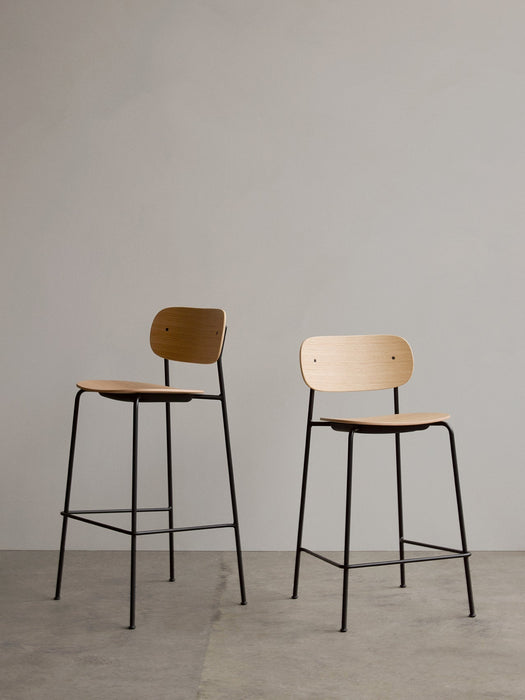 A Co Bar Chair and Co Counter Chair with natural oak veneer seats and black steel frames standing against a plain grey wall.