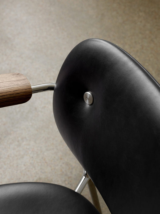 Close-up of the Co Dining Chair's back, featuring black upholstery with a metal stud, a wooden armrest, and a chrome frame.