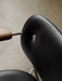 Close-up of the Co Dining Chair's back, featuring black upholstery with a metal stud, a wooden armrest, and a chrome frame.