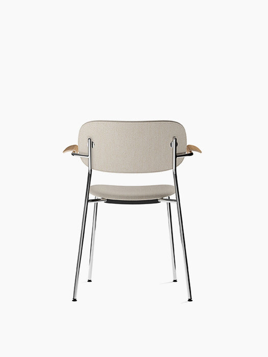 Back of a fully upholstered Co Dining Chair with armrests. It has light fabric, natural oak armrests and a chrome frame.