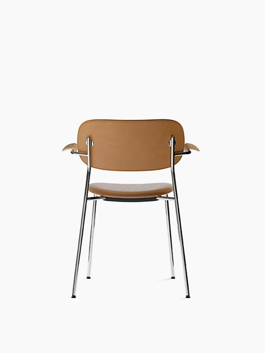 The Co Dining Chair seen from behind, featuring tan Dakar leather upholstery, natural oak armrests, and a slim chrome frame.