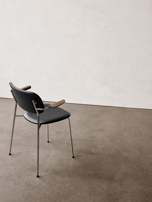 A back three-quarter view of the Co Dining Chair with black upholstery, wooden armrests, chrome frame on a concrete floor.
