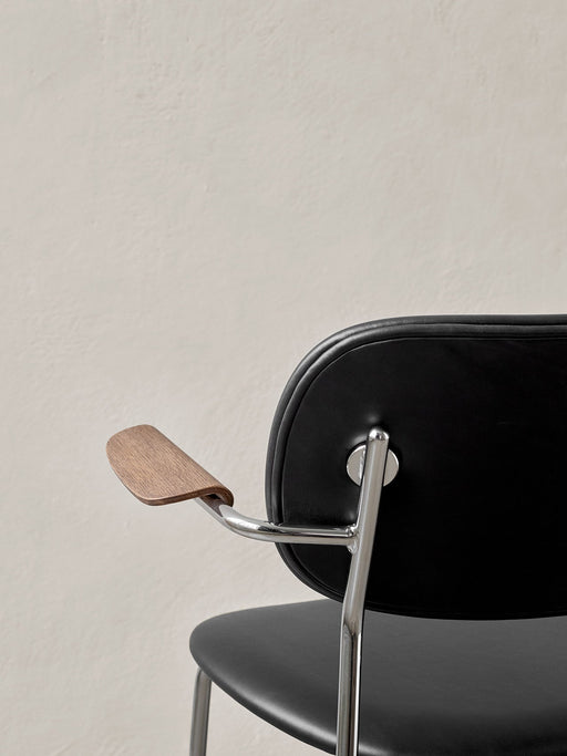 A close-up of the Co Dining Chair's black upholstered backrest, showing the polished chrome frame and the wooden armrest cap.