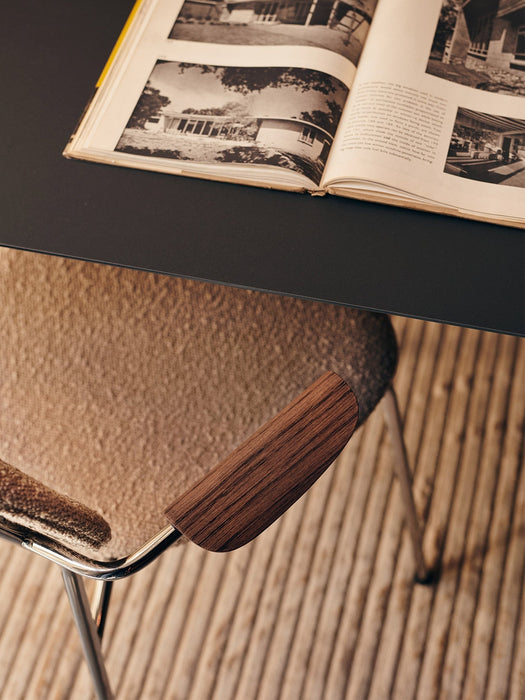 A close-up of the Co Dining Chair's dark wood armrest and beige upholstery, tucked under a black table with an open book.