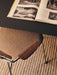 A close-up of the Co Dining Chair's dark wood armrest and beige upholstery, tucked under a black table with an open book.