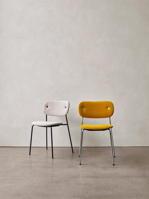 Two fully upholstered Co Dining Chairs. One in off-white bouclé with a black frame, one in yellow with a chrome frame.