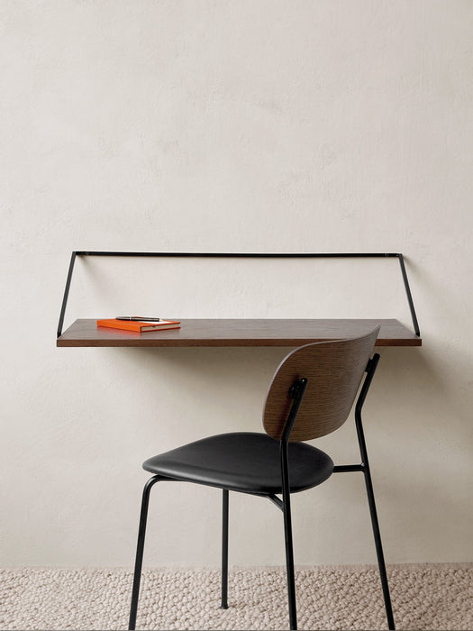 The Co Dining Chair with a black upholstered seat and dark wood back, set at a wall-mounted desk in a minimalist room.