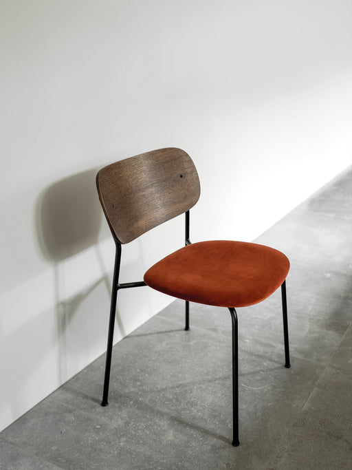A Co Dining Chair with a black frame, dark wood backrest, and orange upholstered seat on a grey floor against a white wall.
