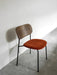 A Co Dining Chair with a black frame, dark wood backrest, and orange upholstered seat on a grey floor against a white wall.