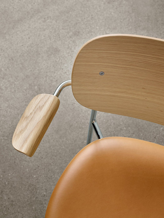 Close-up of the Co Dining Chair with armrest, showing the natural oak veneer back, chrome frame, and tan upholstered seat.