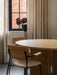 Two Co Dining Chairs with light wood backs and armrests tucked into a large Androgyne Dining Table in front of a window.