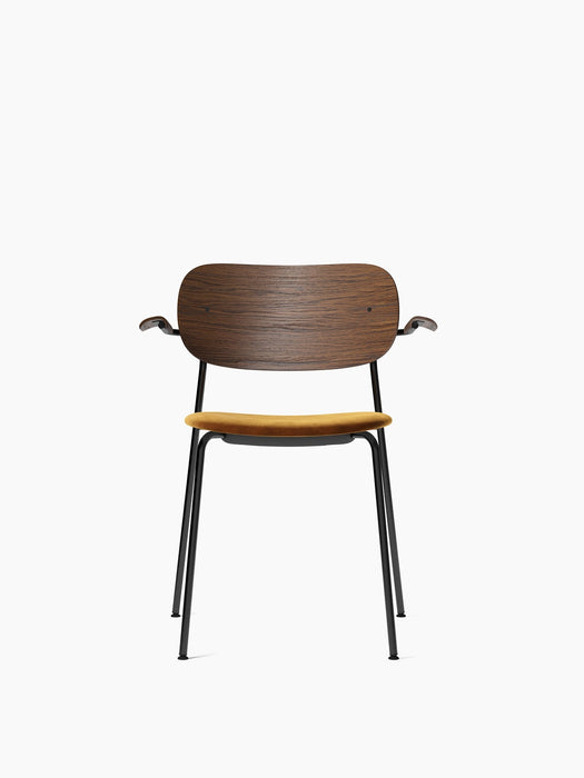 Front view of the Co Dining Chair with armrest, featuring a dark stained oak backrest, mustard seat, and black frame.