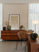 A Co Dining Chair in a stylish room with a wooden sideboard, framed abstract art, a desk, and a glowing floor lamp.
