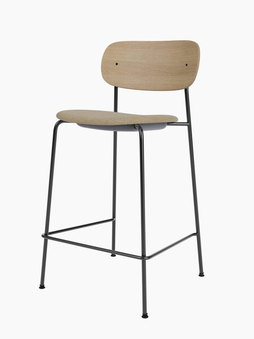 An angled view of the Co Counter Chair with a natural oak backrest, beige upholstered seat, and a slender black steel frame.