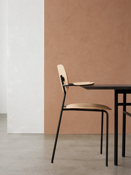 Side profile of the Co Dining Chair with armrest in natural oak veneer, against a minimalist white and terracotta background.