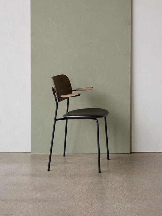 A side profile of the Co Dining Chair with armrests, showing its dark oak back and black frame against a sage green wall.