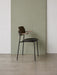 A side profile of the Co Dining Chair with armrests, showing its dark oak back and black frame against a sage green wall.