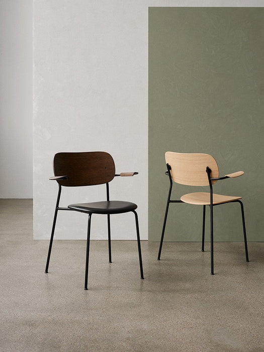 A side-by-side shot of two Co Dining Chairs with armrests: one dark stained oak with a black seat, the other natural oak.