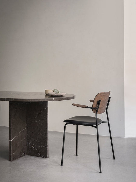 A Co Dining Chair with armrests, featuring a dark wood veneer back and a black seat, sits next to a dark marble dining table.