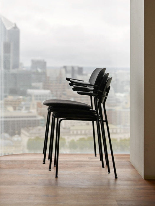 Side view of three black Co Dining Chairs with armrests, stacked on a wooden floor beside a window with a city view.
