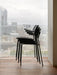 Side view of three black Co Dining Chairs with armrests, stacked on a wooden floor beside a window with a city view.