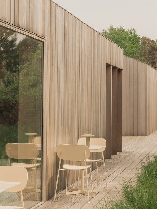 Two ivory Co Dining Chairs, Outdoor and a Nook Dining Table with a Ray Table Lamp on a wooden deck beside a modern building.