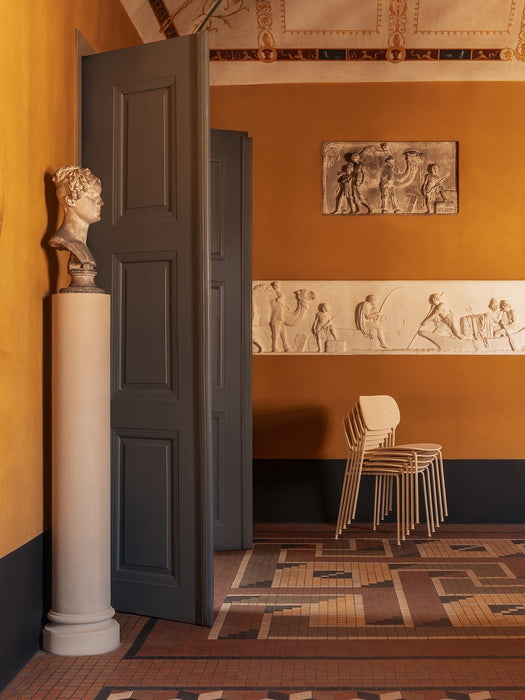 A stack of four ivory Co Dining Chairs, Outdoor, in an elegant, mustard-yellow room with classical bas-reliefs on the wall.