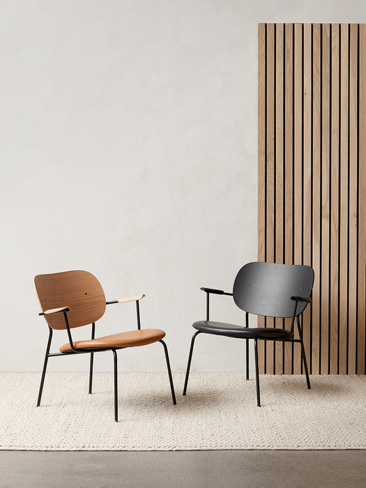 Two Co Lounge Chairs, one in natural oak with a tan seat and one in black, placed on a light rug before a slatted wood panel.