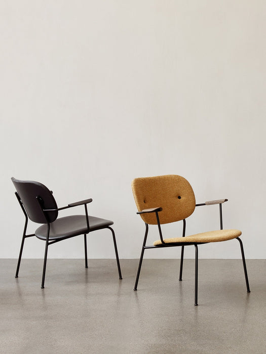 A black Co Lounge Chair and a yellow fabric Co Lounge Chair are shown side by side in a minimalist room with a grey floor.