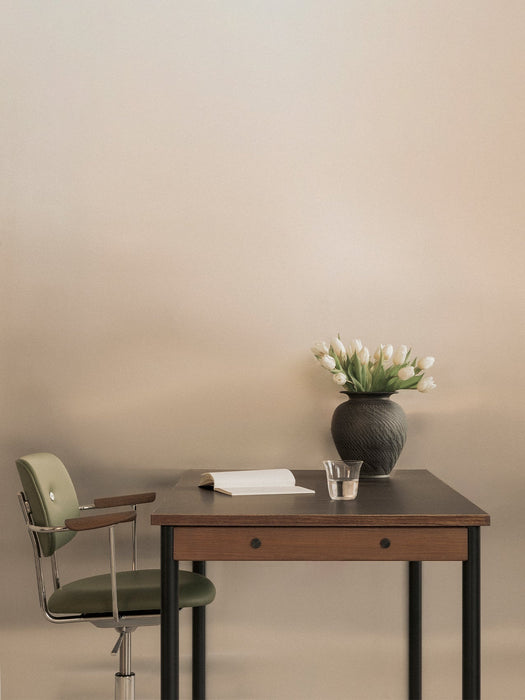 A green Co Task Chair with armrests is pulled up to a dark wood desk with a book, glass of water, and a vase of white tulips.