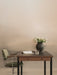 A green Co Task Chair with armrests is pulled up to a dark wood desk with a book, glass of water, and a vase of white tulips.