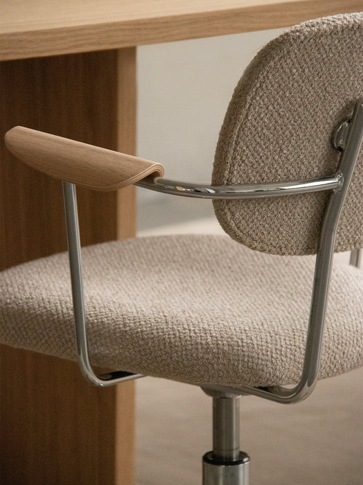 Close-up of the Co Task Chair by a desk, showing its oak armrest, chrome frame, and textured light-coloured upholstery.