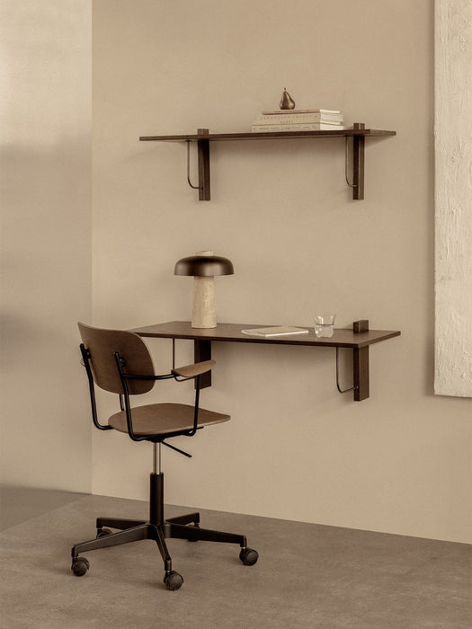 A Co Task Chair at a dark wood Corbel Desk with a Reverse Table Lamp. Above, a Corbel Shelf holds books and a pear.