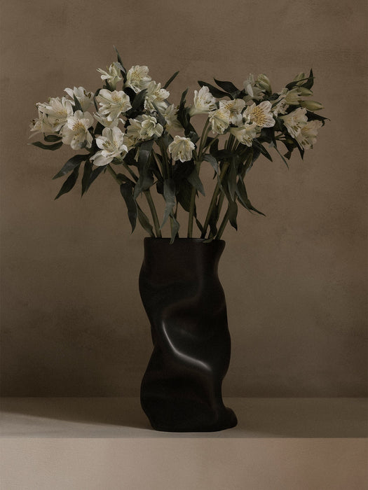 The black, sculptural Audo Collapse Vase sits on a light surface, holding a bouquet of white flowers against a taupe wall.